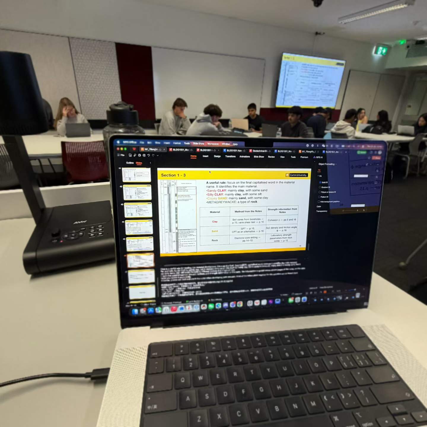 The BLDG1001 classroom seen from behind the teaching laptop, with students working at their tables.